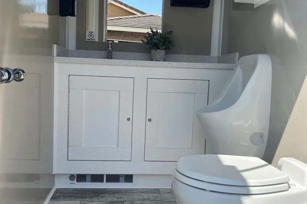 Interior of a white portable restroom, featuring a sink, cabinets, urinal, and toilet.