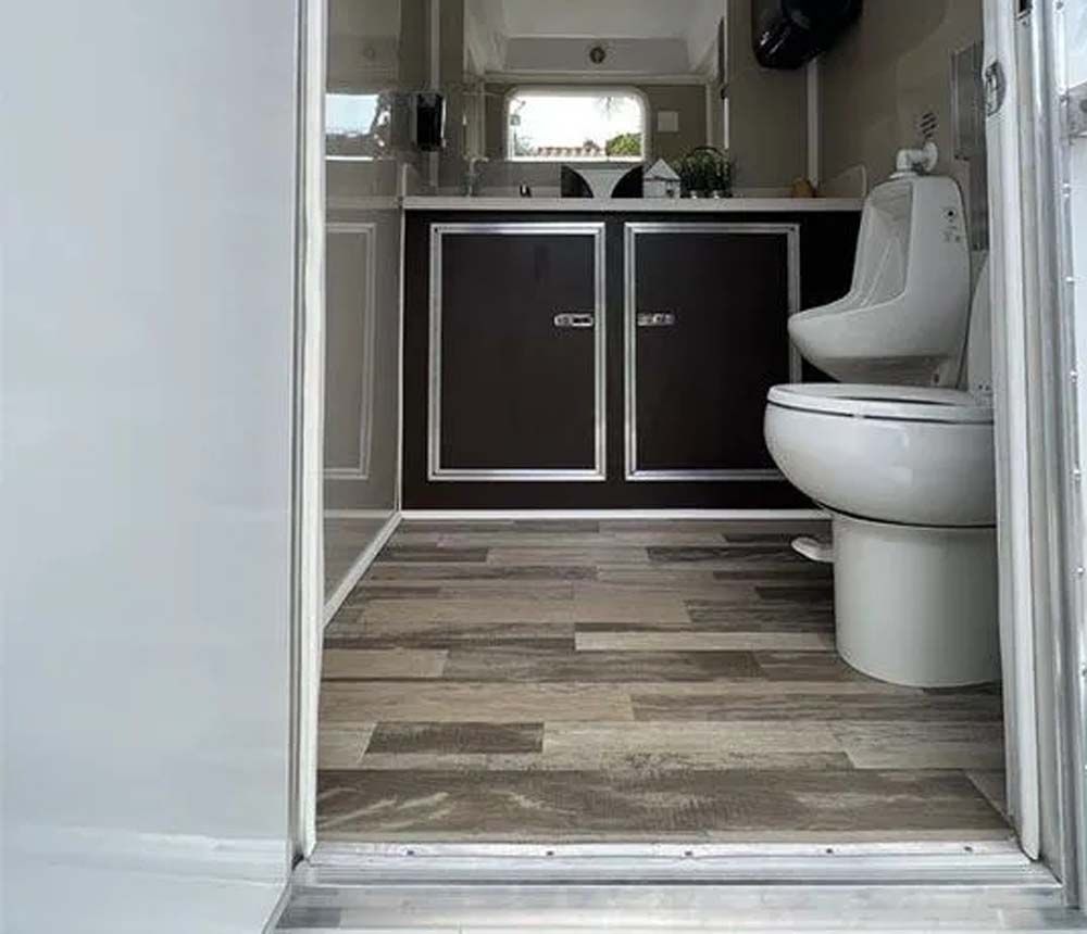 Bathroom trailer interior with toilet, urinal, and sink. Wood-look floor, silver and brown accents.