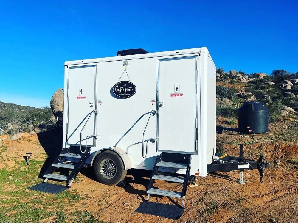 White portable restroom trailer on a trailer, with steps. Set in a natural outdoor area.