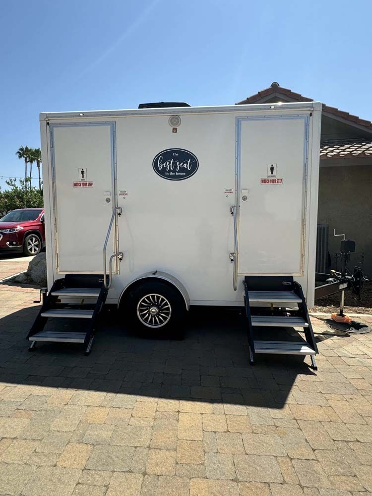 White portable restroom trailer with two doors and steps, parked outside.