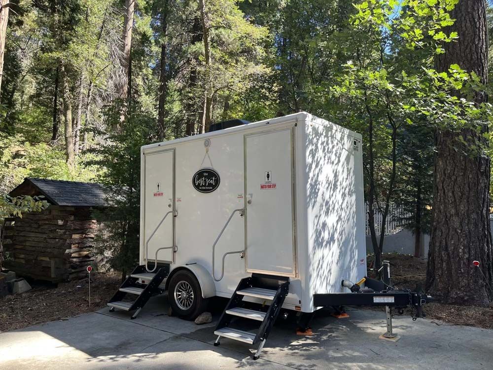 White portable restroom trailer in a wooded area, with steps and a trailer hitch.