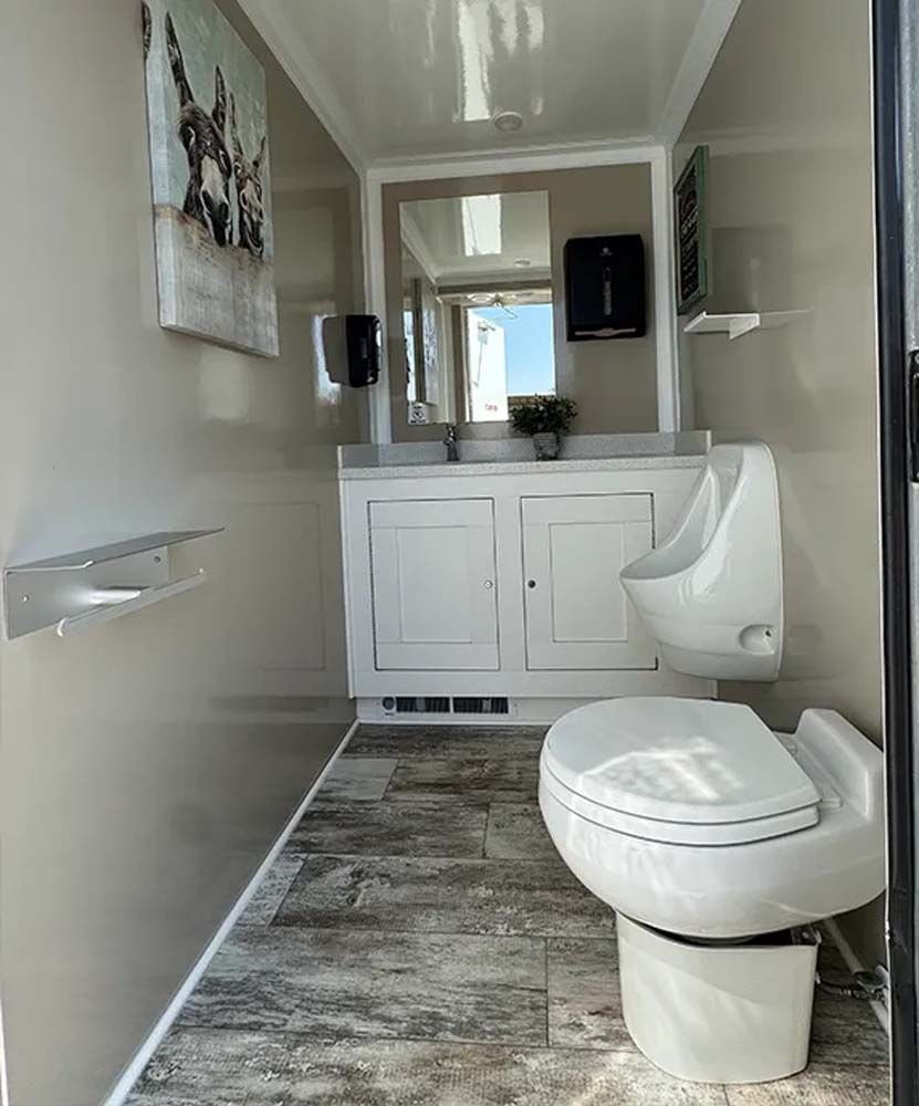 Interior of a luxury portable restroom with toilet, urinal, sink, and artwork.