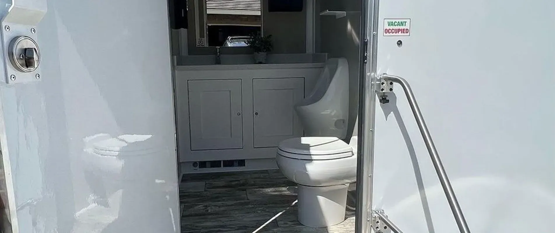 Open door of a white, portable restroom, revealing a toilet, sink, and mirror.