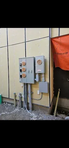 Electrical meters and service panels mounted on an exterior wall with conduit pipes, near a construction area.