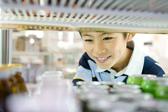 A young boy is looking through a refrigerator filled with bottles and cans.