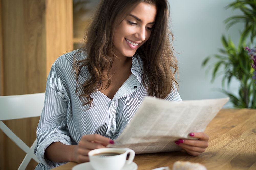 A woman is sitting at a table reading a newspaper and drinking coffee.