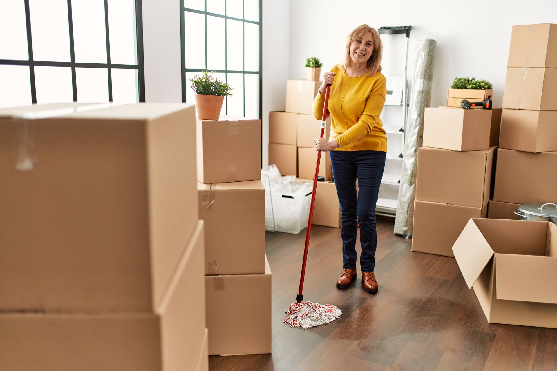 Woman mopping a room filled with moving boxes. She smiles, wearing a yellow sweater and jeans.