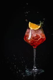 Un bicchiere di liquido rosso con ghiaccio e una fetta d'arancia su uno sfondo nero.