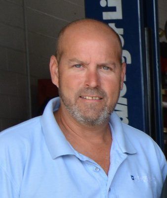 Man with a shaved head and graying beard, wearing a light blue polo shirt, standing in a garage. | Heaven Sent Automotive, LLC