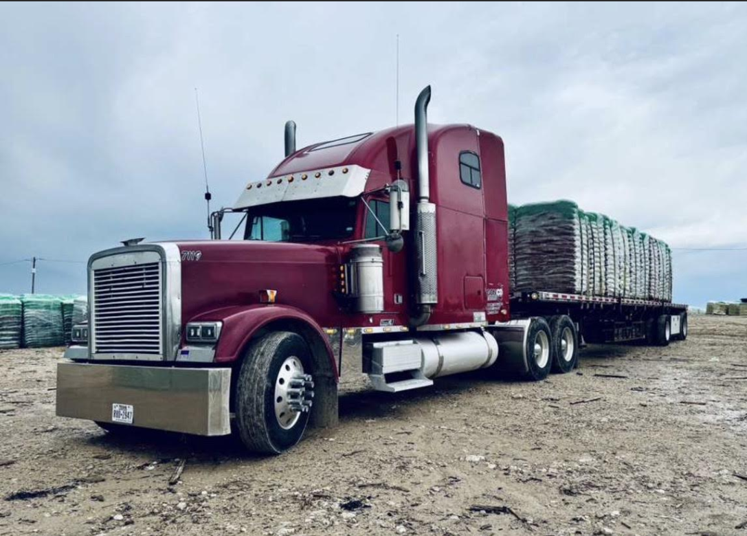 Maroon semi truck hauling a long flatbed load on a gravel lot under a cloudy sky