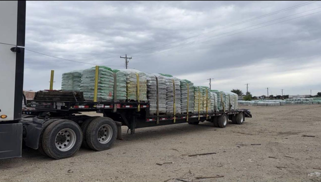 Flatbed truck loaded with stacked pallets driving across a gravel lot under cloudy skies