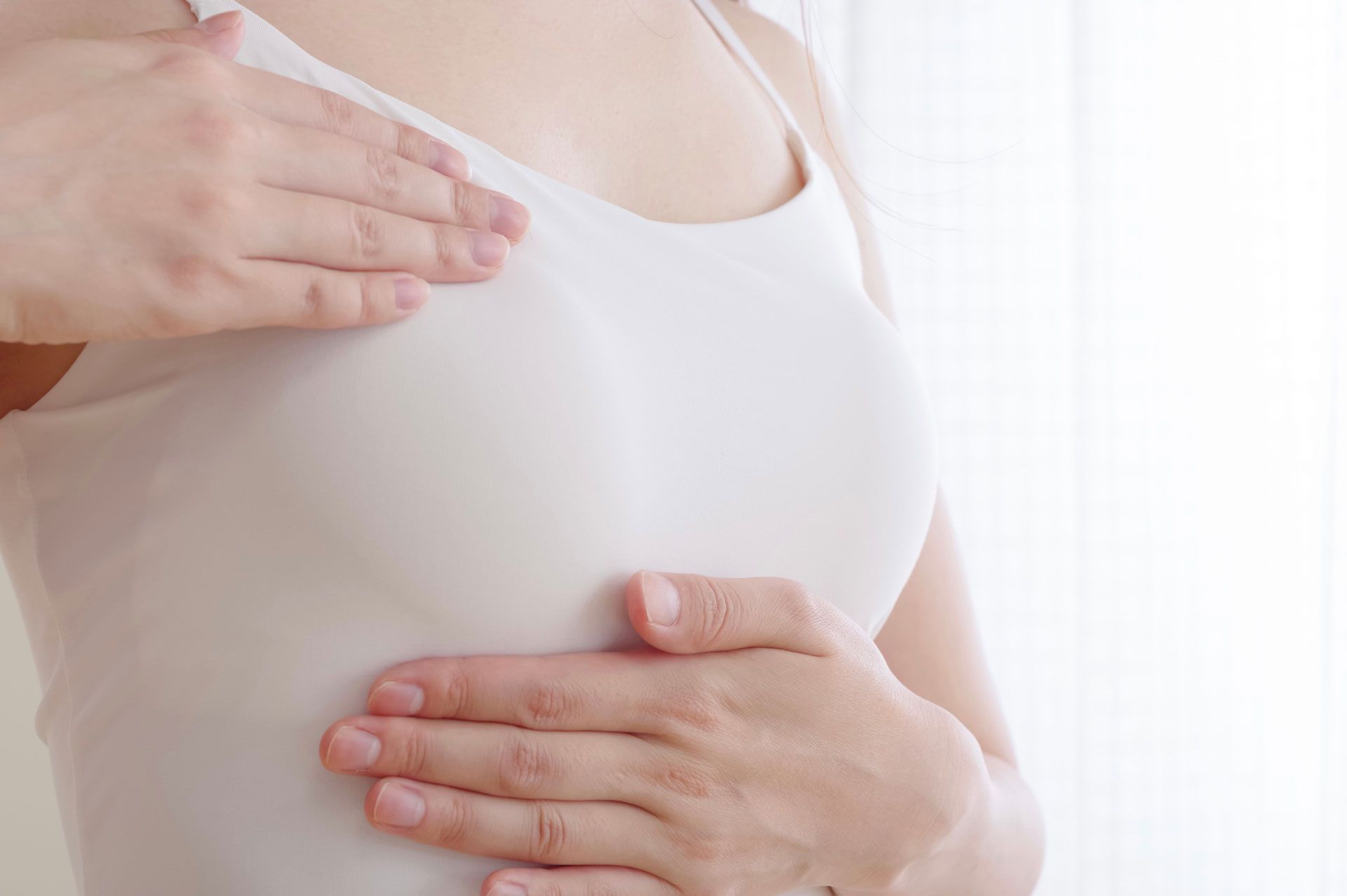A person’s hands performing a self-examination on their chest while wearing a white tank top.
