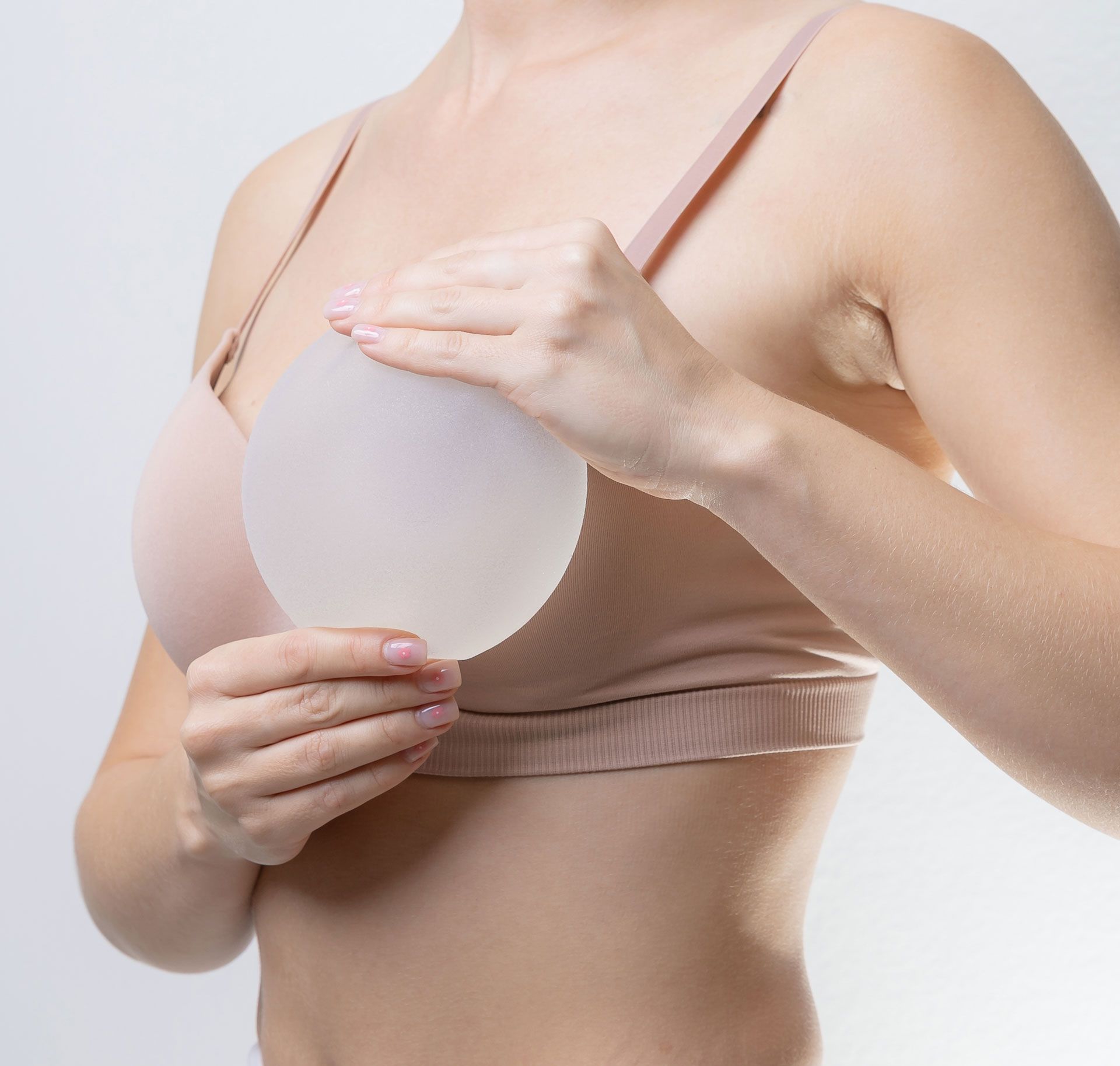 A person in a beige bra holds a round, translucent silicone breast implant in front of their chest.