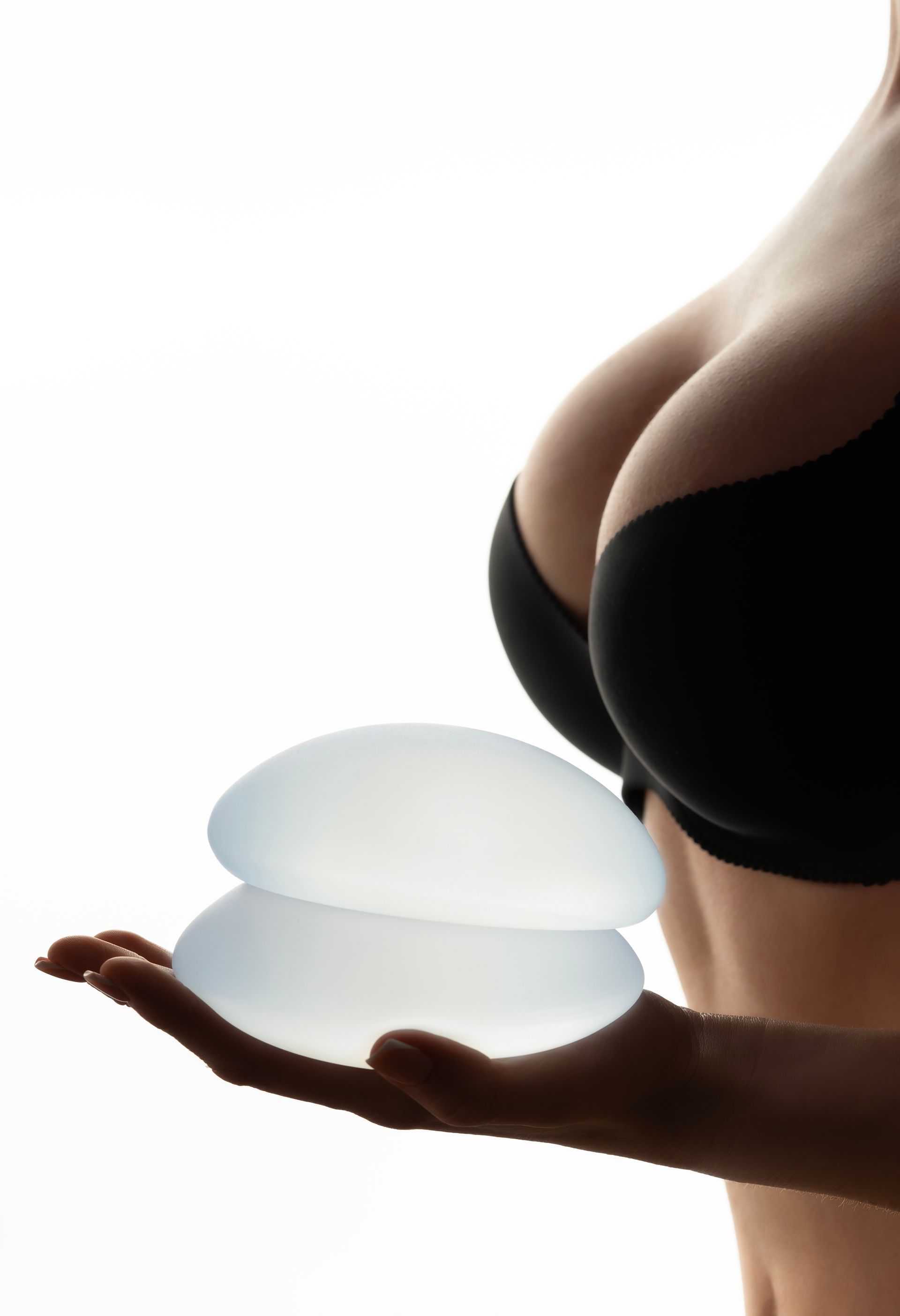A person in a beige bra holds a round, translucent silicone breast implant in front of their chest.