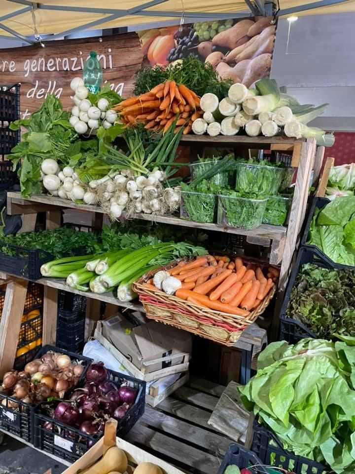 Bancarella di verdura al mercato, che espone carote, cipolle, lattuga e altri prodotti freschi.