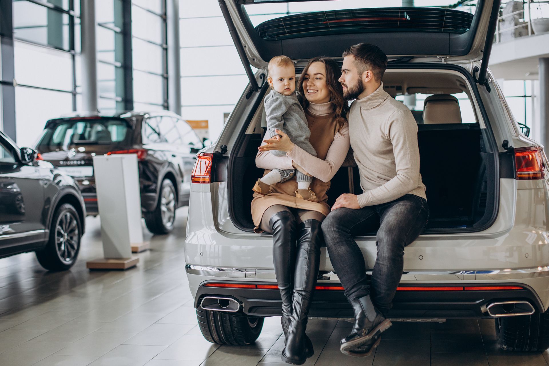 Family of three sitting in the open trunk of a silver SUV at a car dealership.