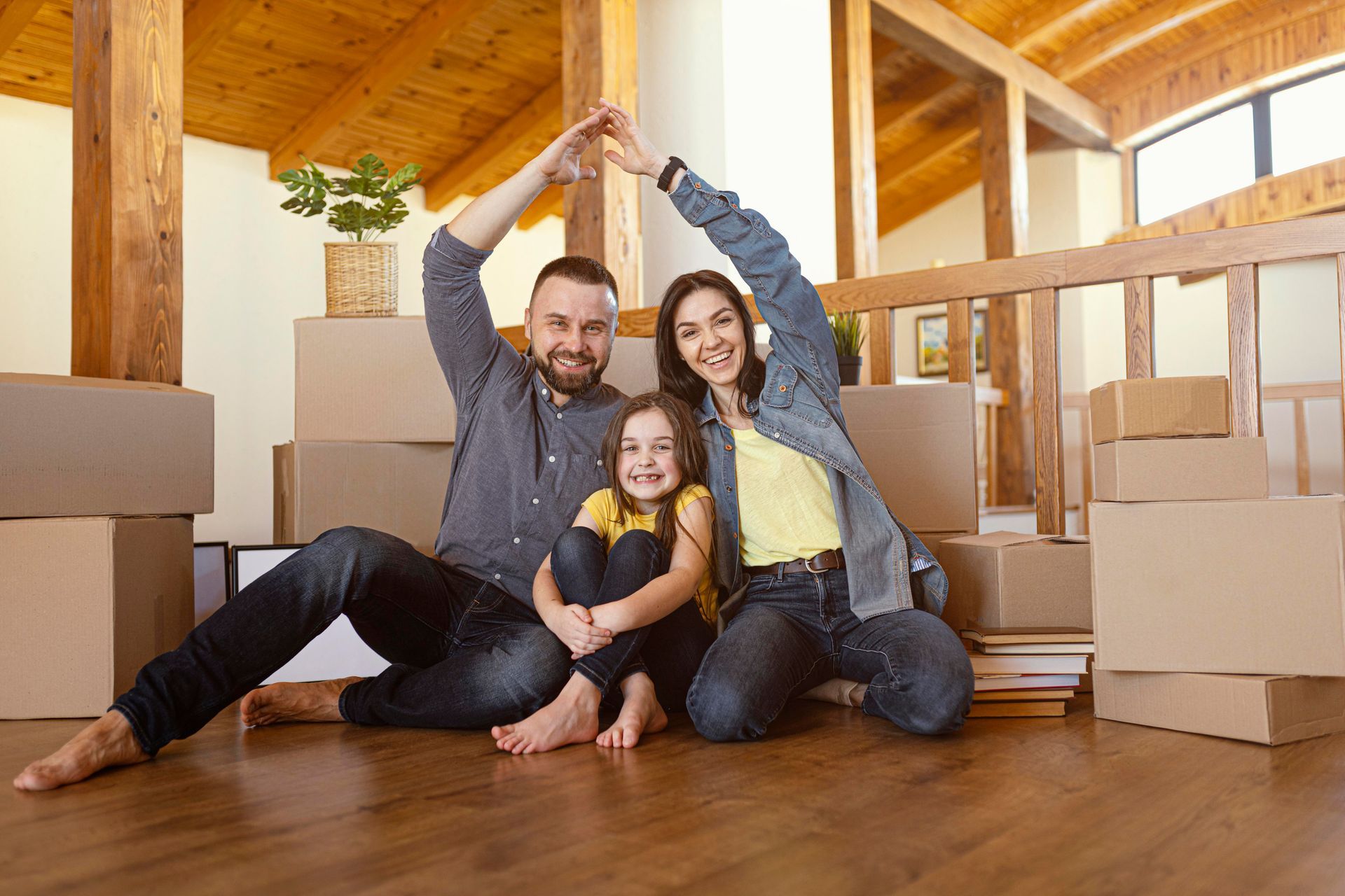 Family of three making a house shape with arms, surrounded by moving boxes, smiling happily.