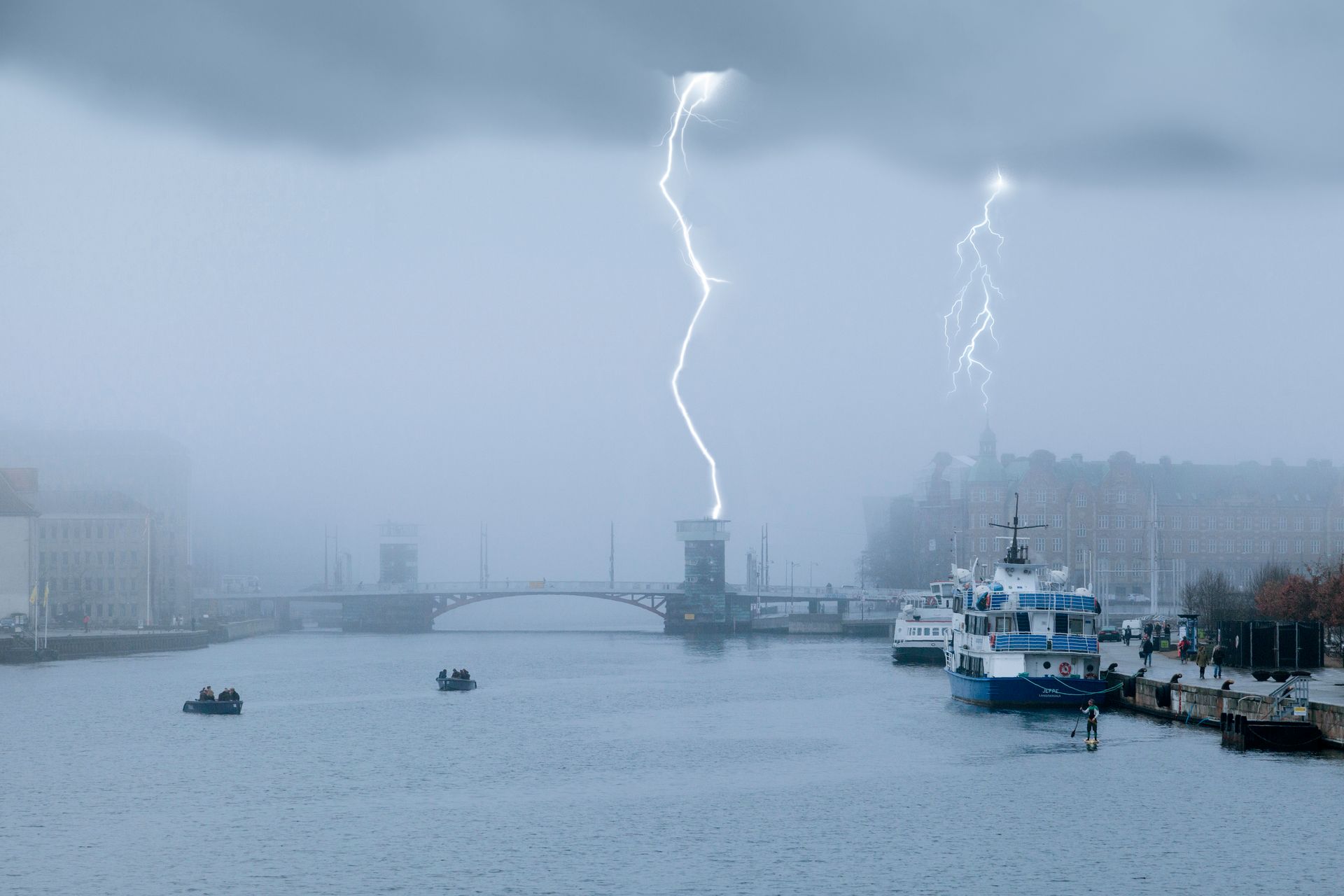 Lightning strikes over a bridge and boats on a foggy river.