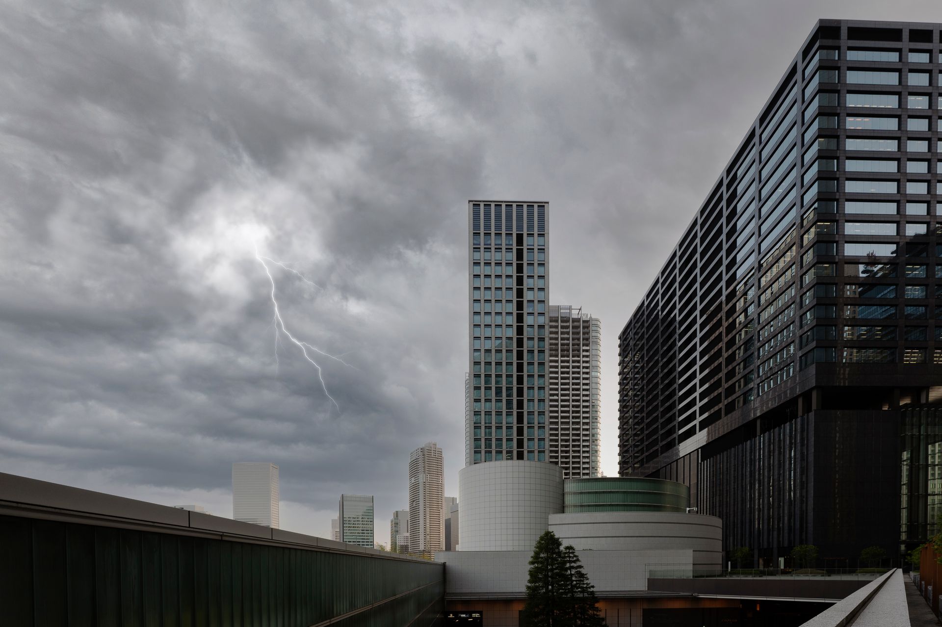 Lightning strikes over a modern cityscape under a cloudy, gray sky. Tall buildings stand in the background.