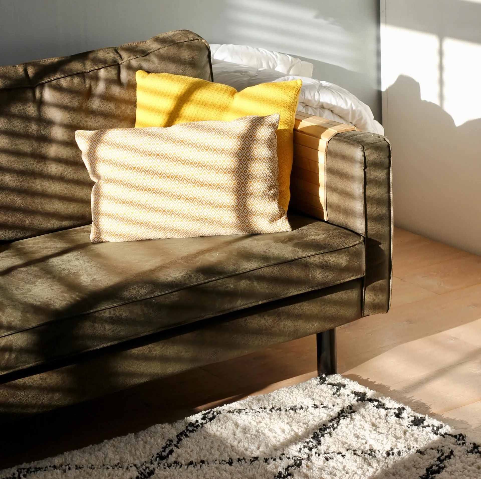 Brown sofa with yellow and cream pillows, lit by sunlight through blinds; rug on floor.