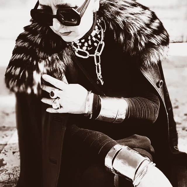 Persona seduta all'aperto con occhiali da sole, collo di pelliccia e gioielli d'argento.