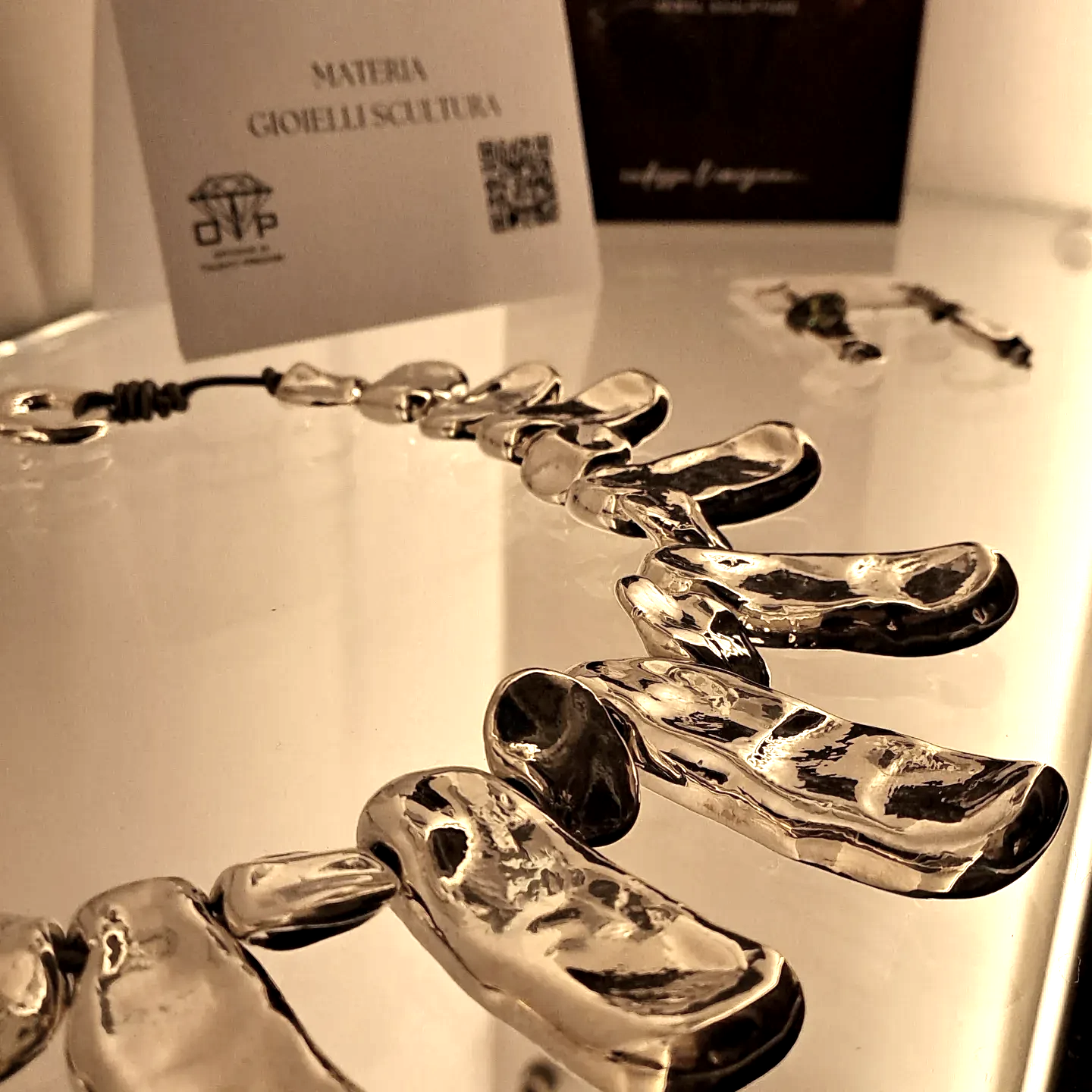 Collana scultorea in argento su una superficie riflettente con l'etichetta del prodotto sullo sfondo.