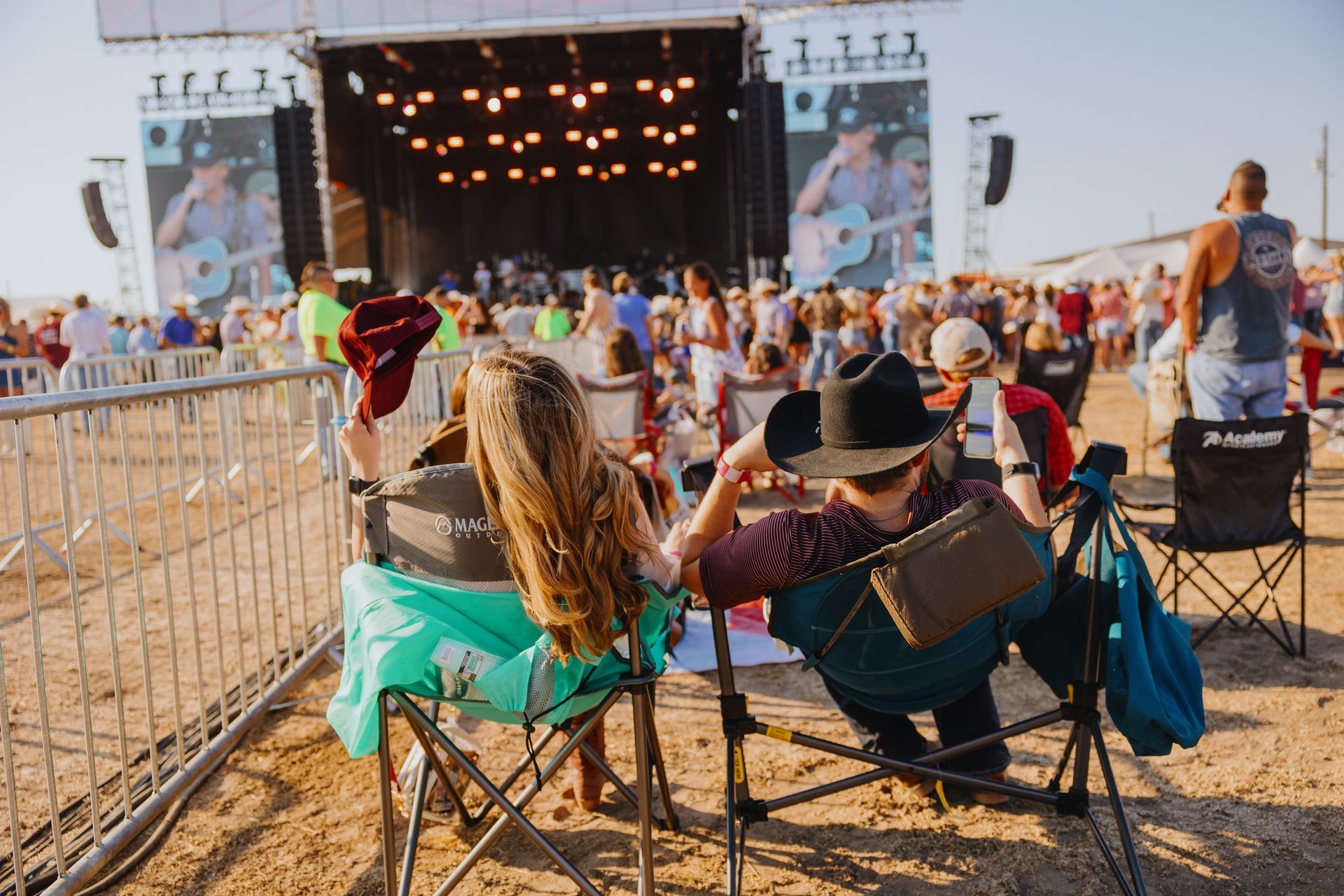 Two women sit in chairs at a concert, looking at the stage. Many people stand near a fence.