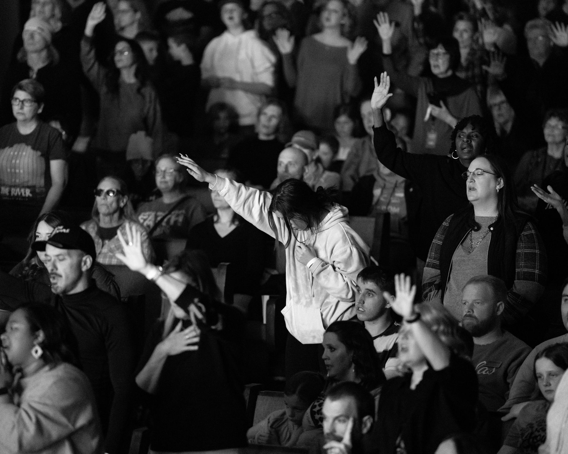People raising their hands in a crowd, some with open mouths, possibly singing or worshipping in a dark setting.