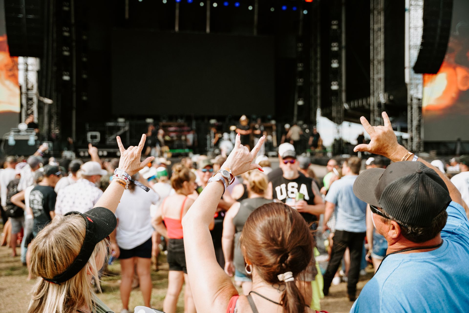 Crowd at outdoor concert, people raising hand gestures, stage with screen in background, sunny day.