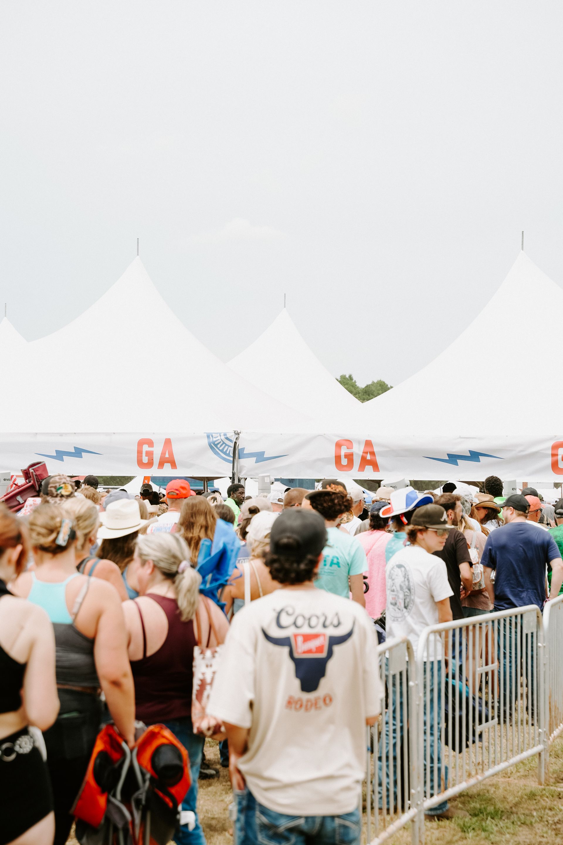 People waiting in line at a festival entrance, under white tents.
