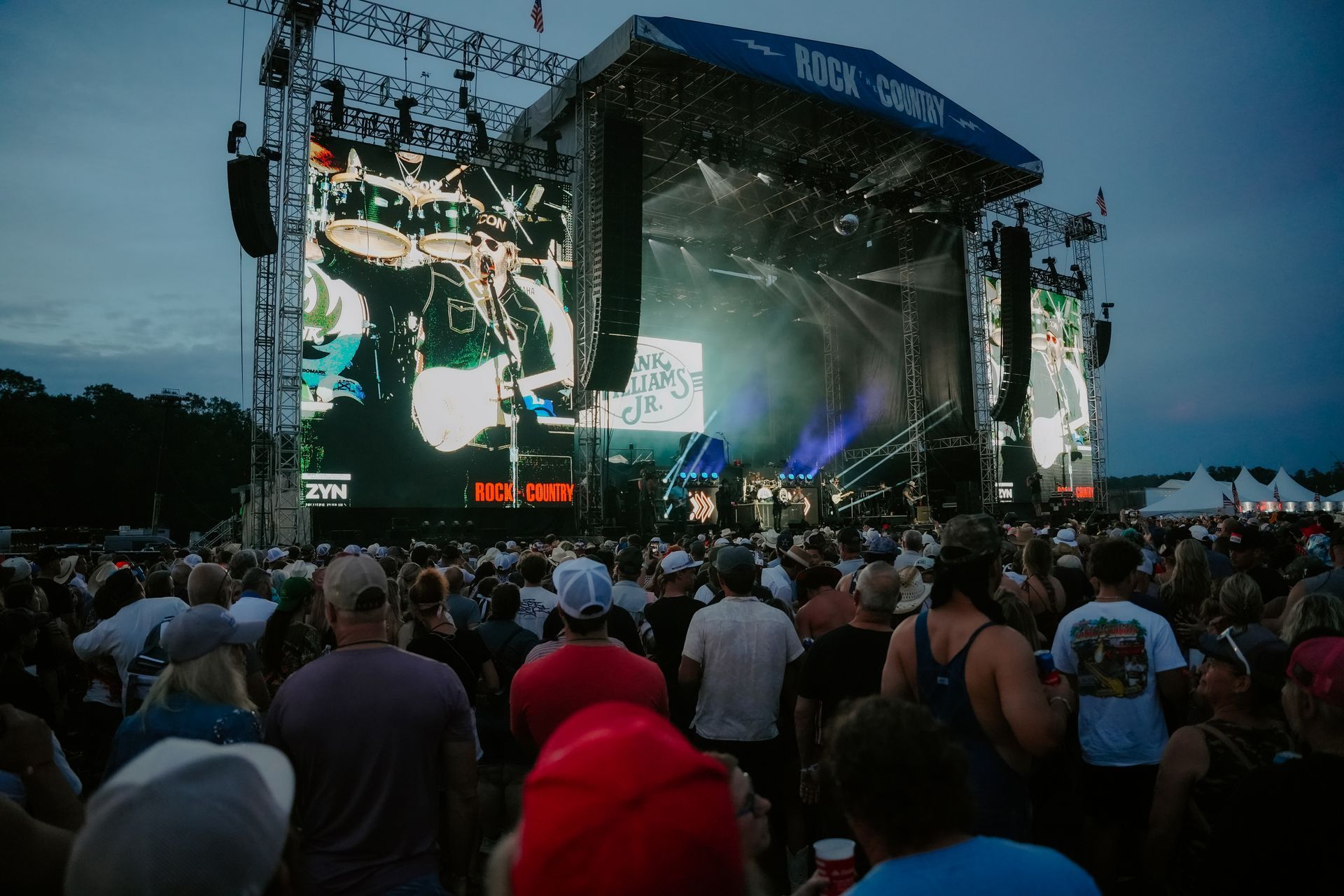 Crowd watches band perform on large outdoor stage with screens under a dusky sky.