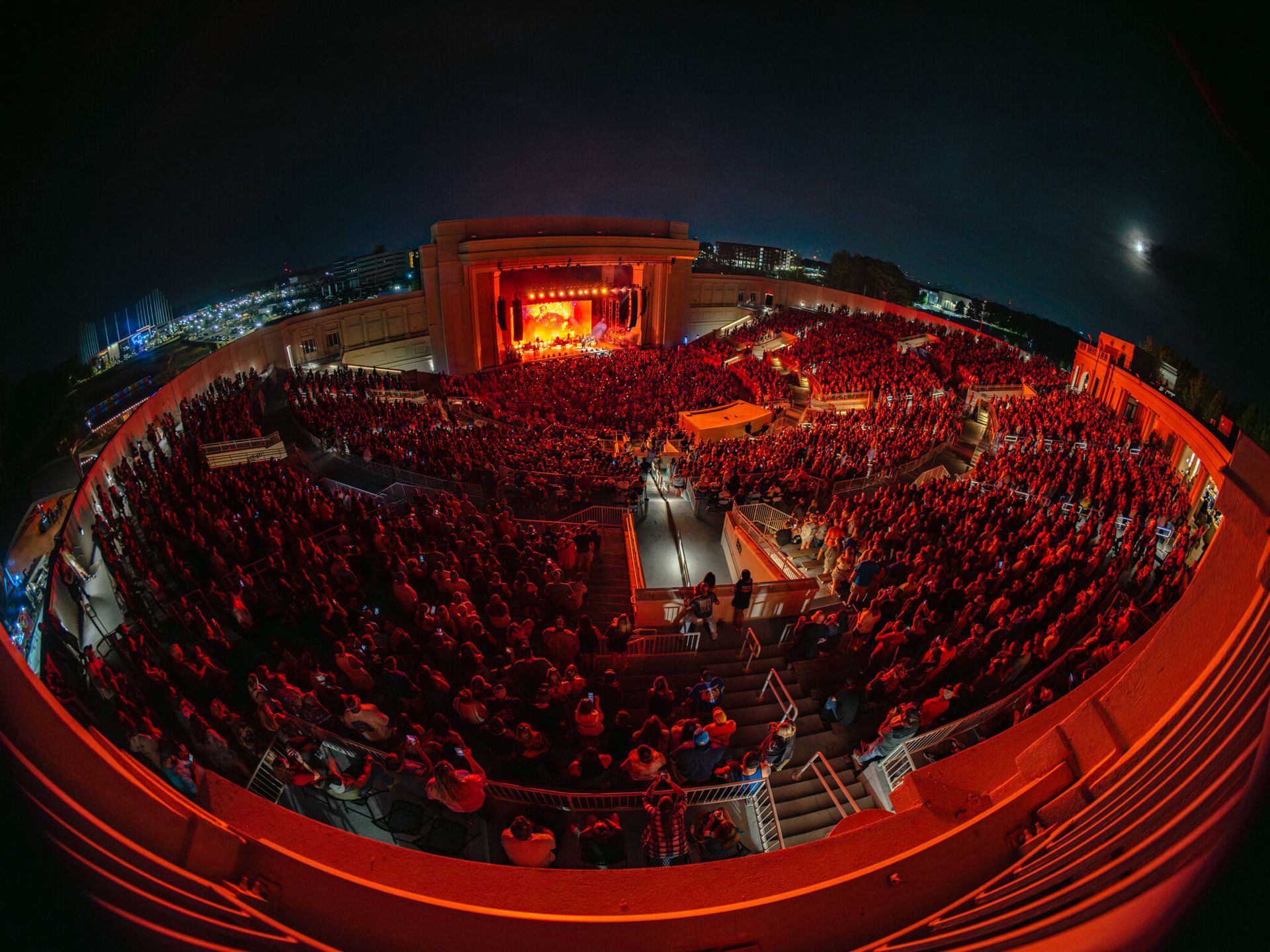 Nighttime concert in an outdoor amphitheater; red stage lights illuminate the crowd and performers.