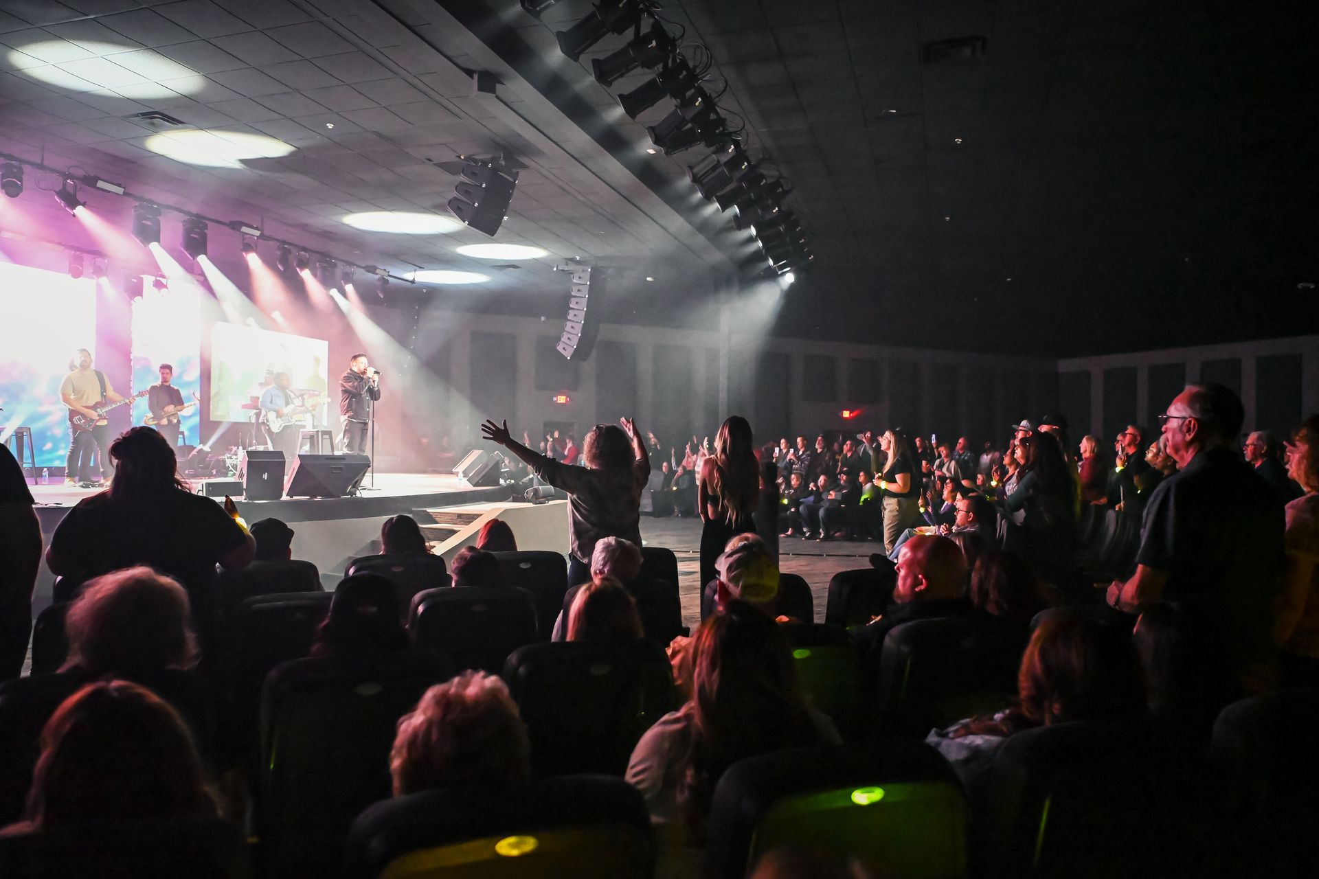 Concert hall: band on stage with spotlights; audience watches, some raise arms, in dark auditorium.