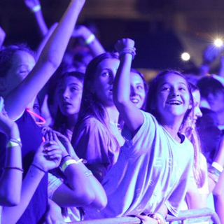 A group of young people at a concert, lit by blue lights, cheering with raised arms and happy expressions.
