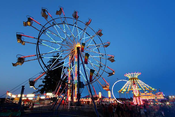Ferris wheel and carnival rides at dusk, illuminated with colorful lights against a blue sky.