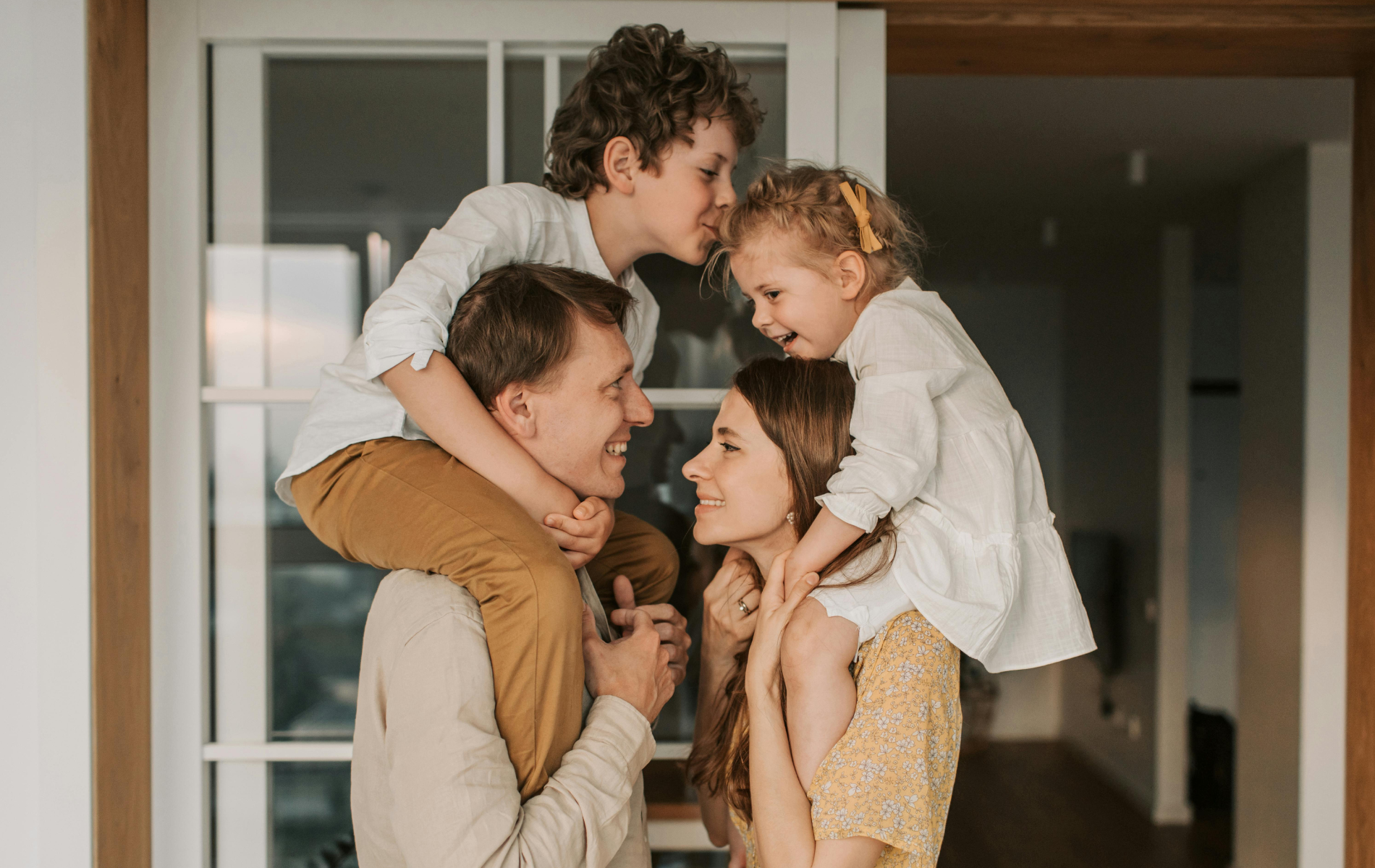 A man and woman each carry a child on their shoulders inside a home, looking at each other with warm expressions.