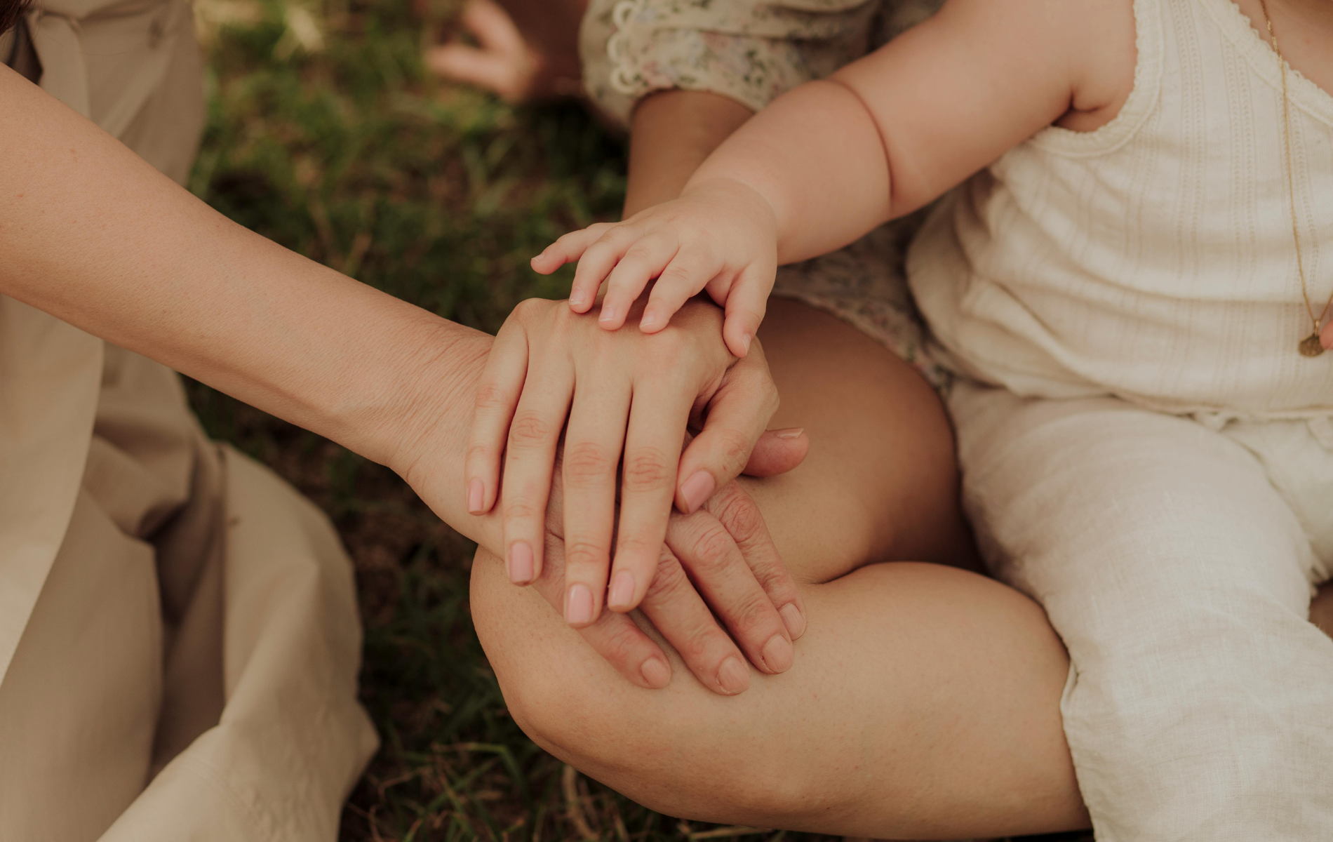 Hands of an adult and a small child stacked together while resting on a lap outdoors.