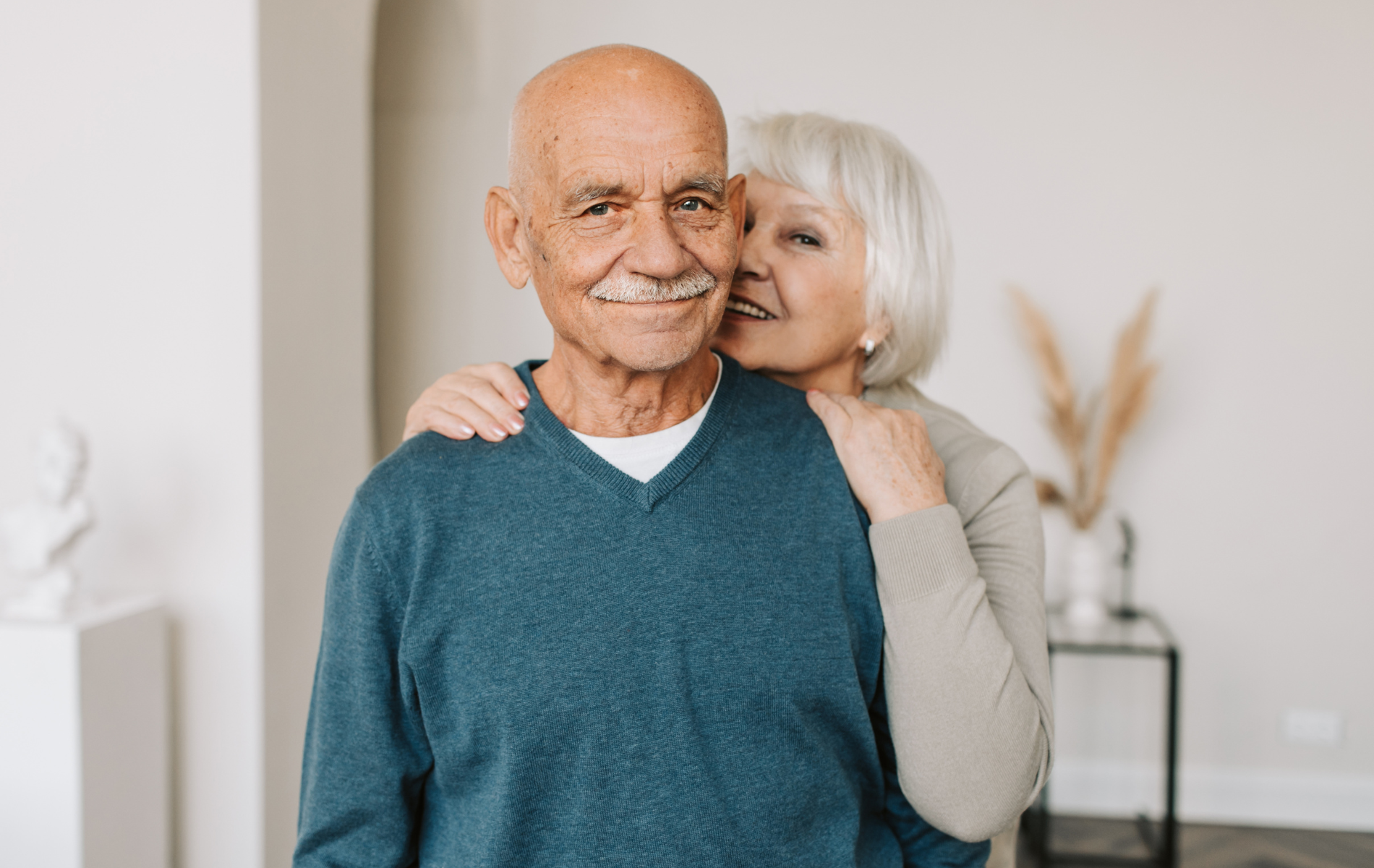 A couple embracing in an indoor space. The person in front wears a blue sweater, and the person behind wears a beige top.