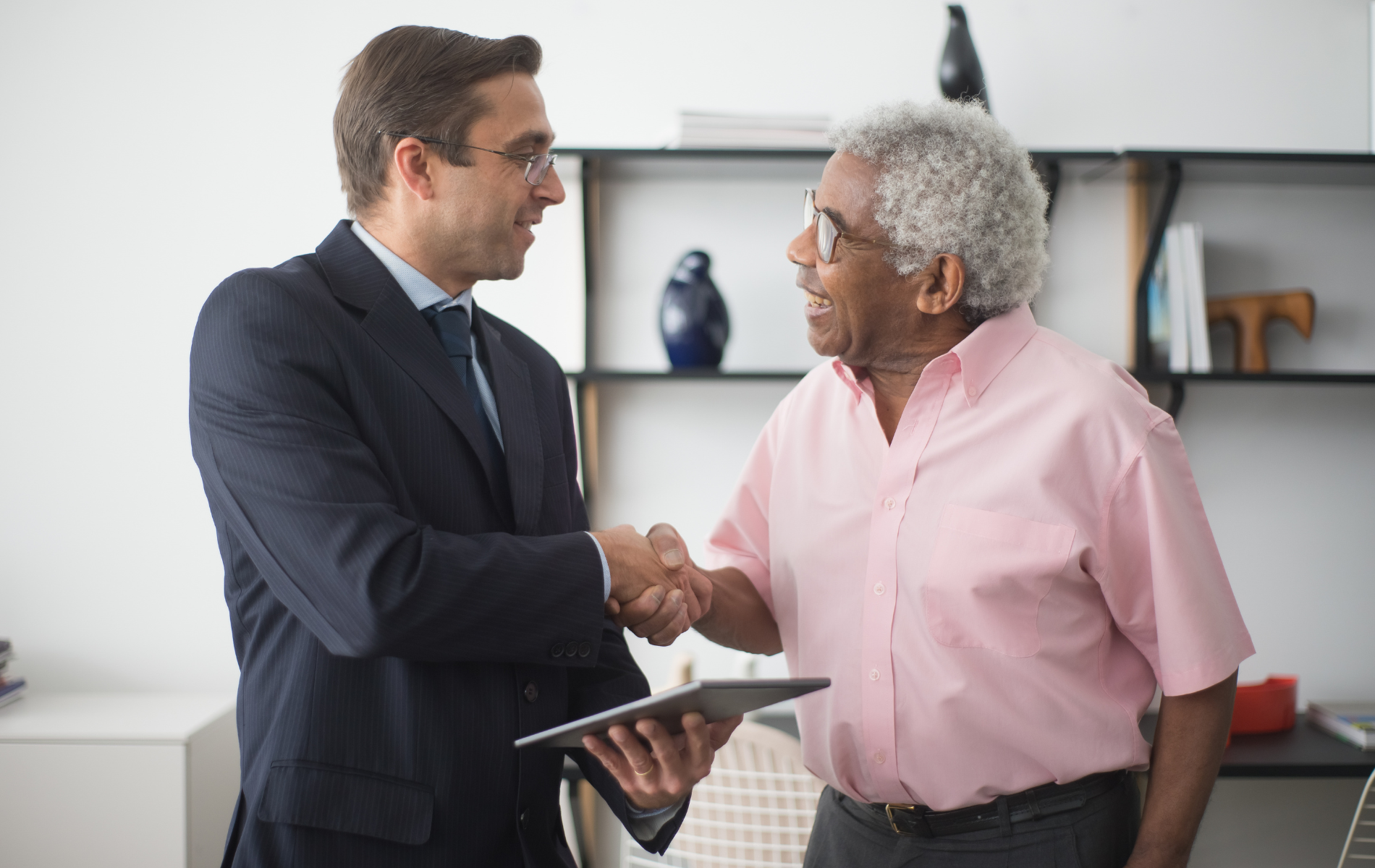Two professionals shaking hands in an office setting while holding a tablet.