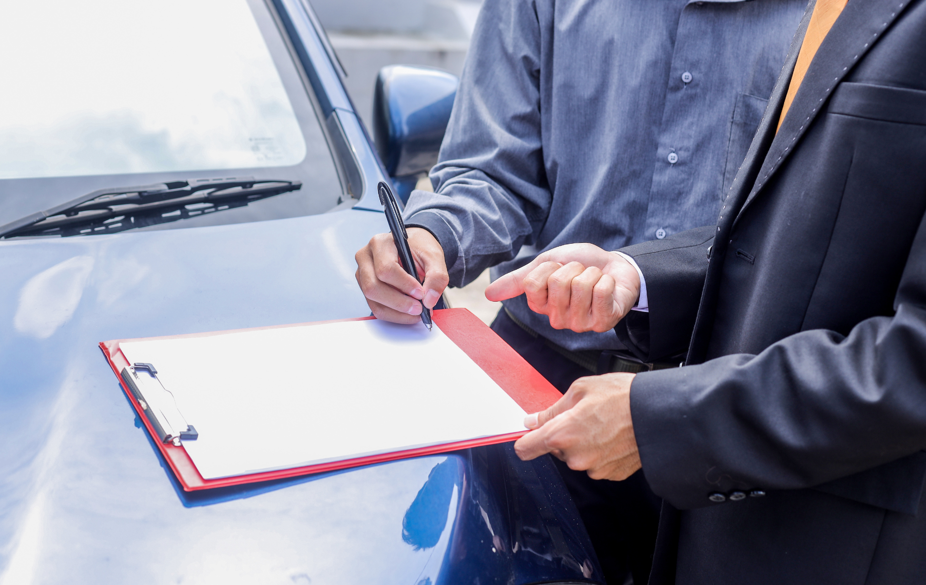 Two people, one in a dark suit and one in a grey shirt, look at a clipboard held over the hood of a blue car.