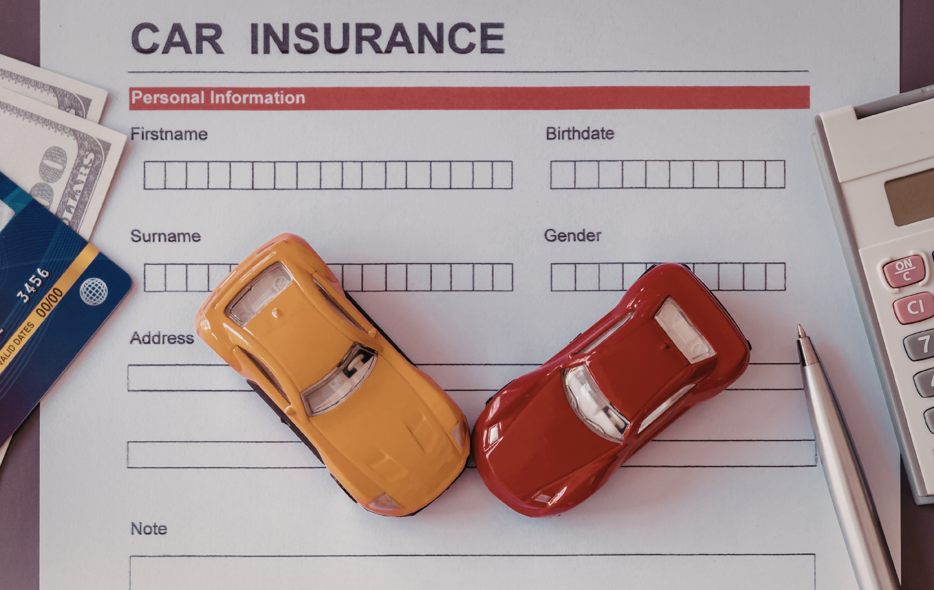 A car insurance form with two toy cars, a calculator, a pen, and money on a surface.