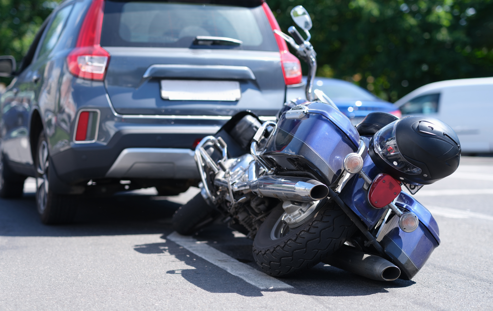 A blue motorcycle lies on its side on the pavement behind a gray SUV parked on a city street.