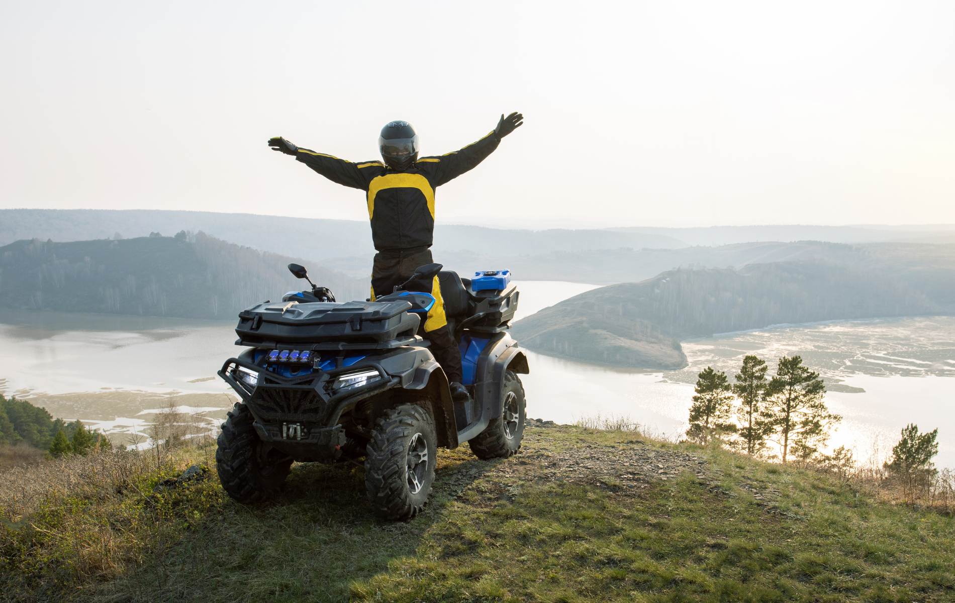 A person in riding gear stands on an ATV with arms raised in victory, overlooking a scenic river valley at sunset.