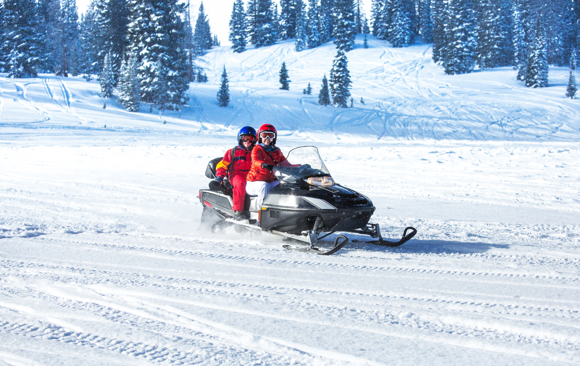 Two people in helmets ride a snowmobile across a snowy, sunlit landscape with pine trees in the background.