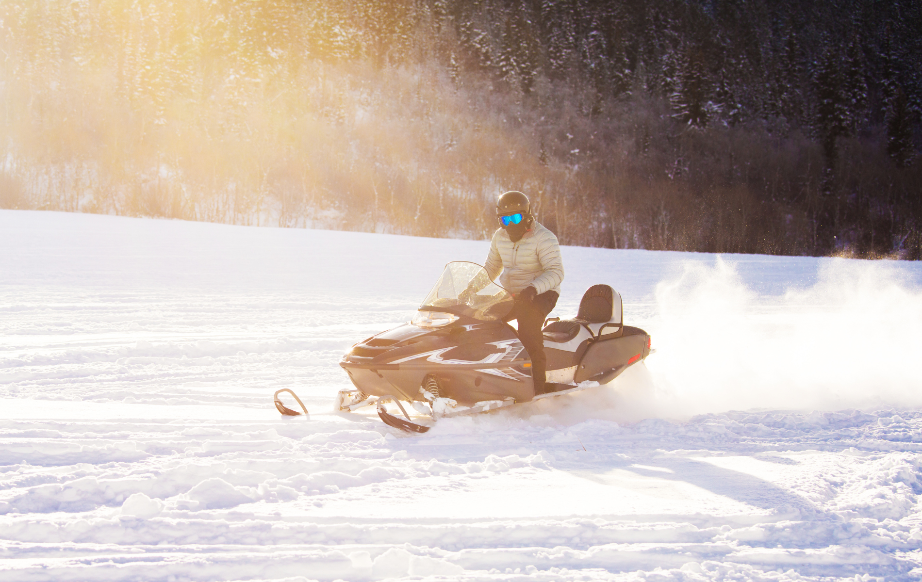 A person wearing a helmet and goggles rides a snowmobile across a sunny, snowy landscape, kicking up powder.