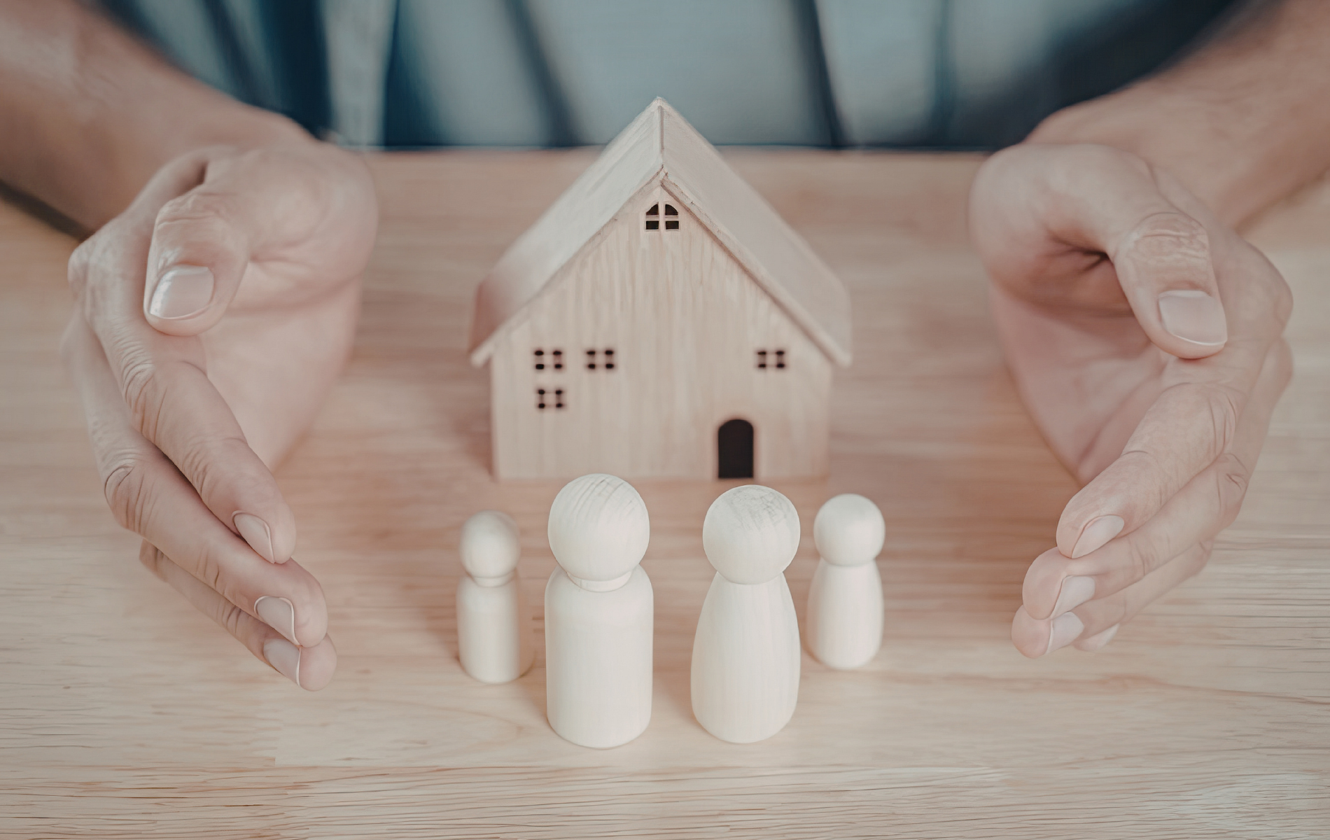 Hands cupping a small wooden model house with four wooden peg dolls standing in front of it on a wooden surface.