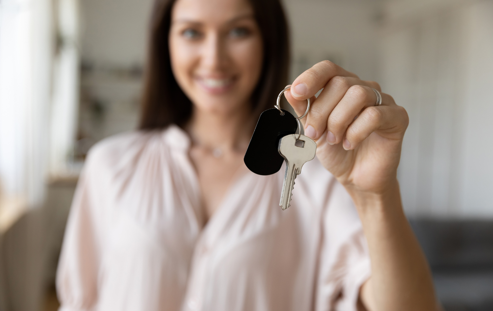 A smiling person holds a key ring featuring a black tag toward the camera, symbolizing a new home or property ownership.