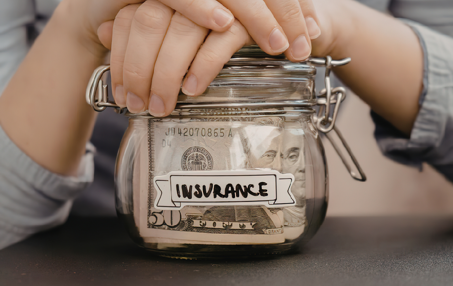 A person’s hands rest on a glass jar filled with US dollar bills, labeled with a white sticker that says 