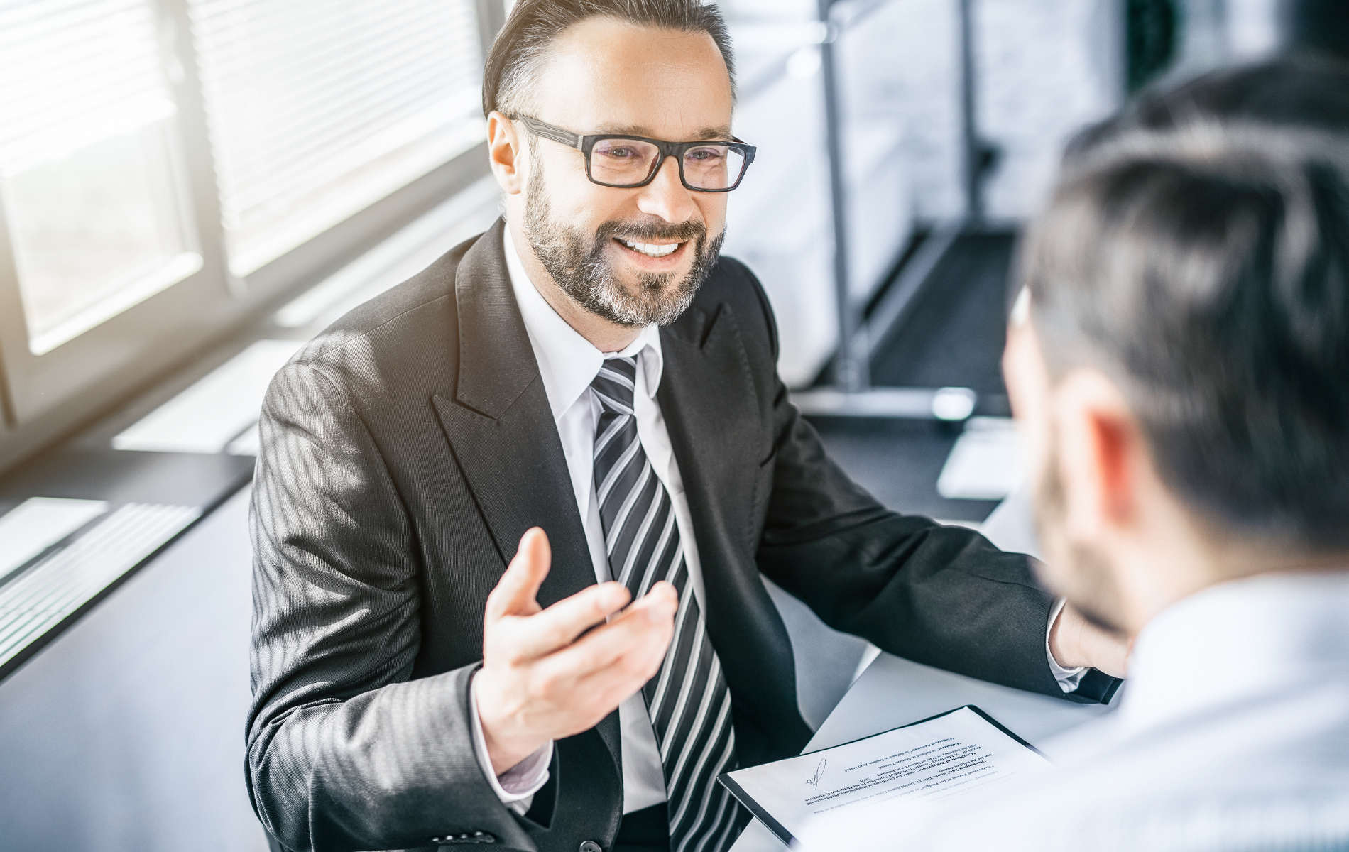 A smiling man in a suit and glasses gestures while speaking to another person in a bright, modern office.