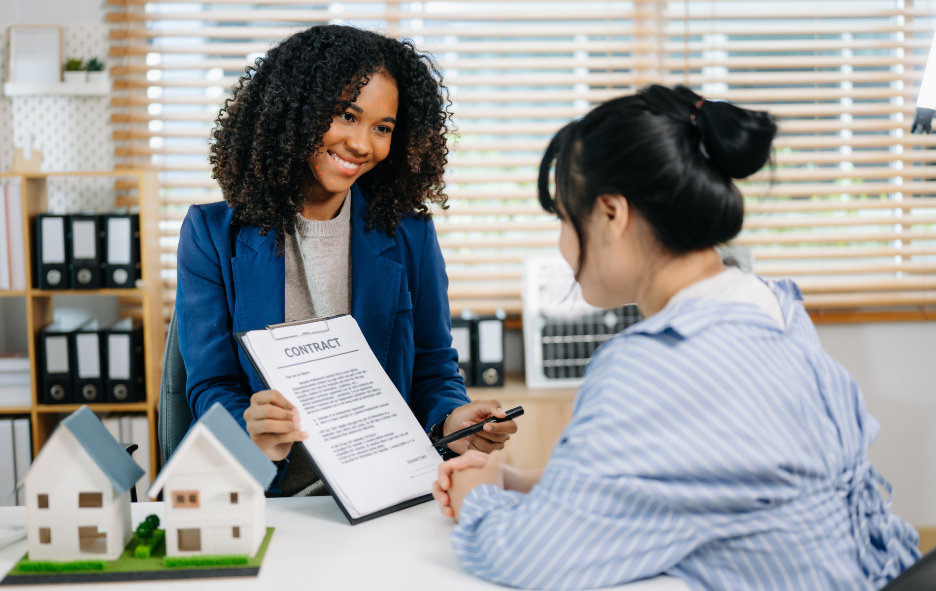 A professional in a blue blazer presents a contract to a client at a desk with two small model houses.